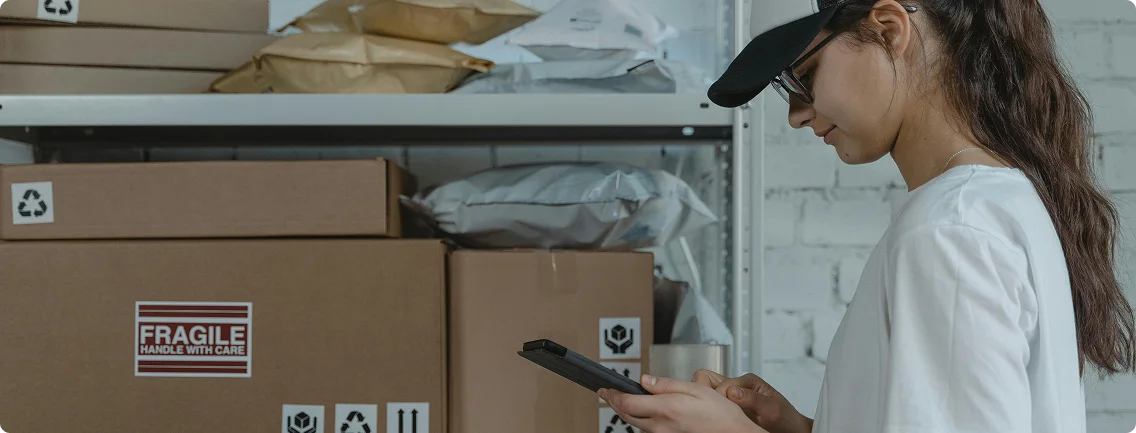 A girl wearing a black cap and glasses, with dark hair, is shown in profile. They are looking down and interacting with a tablet or mobile device held in their hands. Behind them are shelves stacked with various packages and cardboard boxes, some labeled "FRAGILE HANDLE WITH CARE."