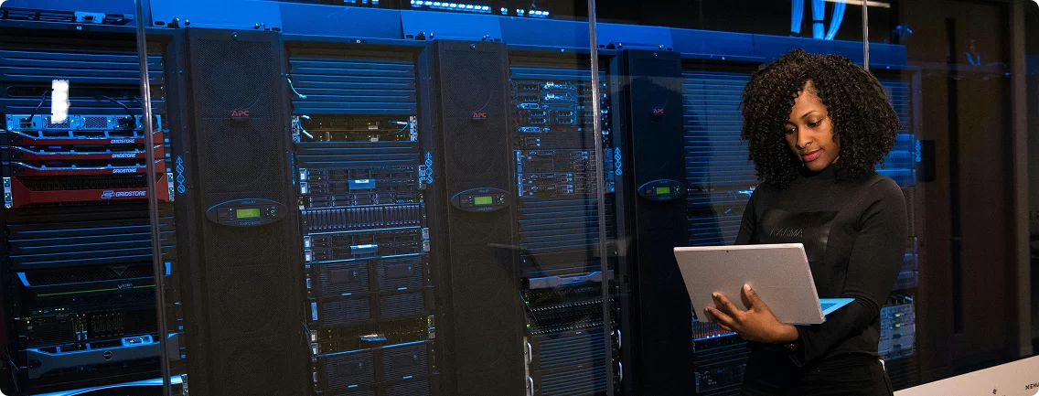 A woman with dark, curly hair stands in front of tall server racks, illuminated by blue lights. She is wearing a dark top and looking down at a silver tablet she holds in both hands, appearing to be working or monitoring the equipment. The server racks dominate the left side of the frame, filled with glowing indicators and components.