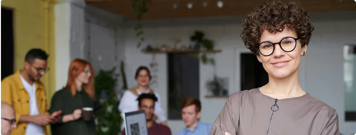 A girl with short, curly brown hair and round glasses smiles confidently at the camera, with their arms crossed. They are wearing a collared, short-sleeved top with a zipper detail. In the blurred background, several other people are visible in what appears to be an office or collaborative workspace, some standing and talking, others seated at a table.