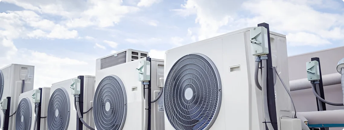 A row of large, white outdoor HVAC condenser units with circular fans is shown under a partly cloudy sky. The units are lined up on what appears to be a rooftop, with pipes and electrical conduits connecting them.